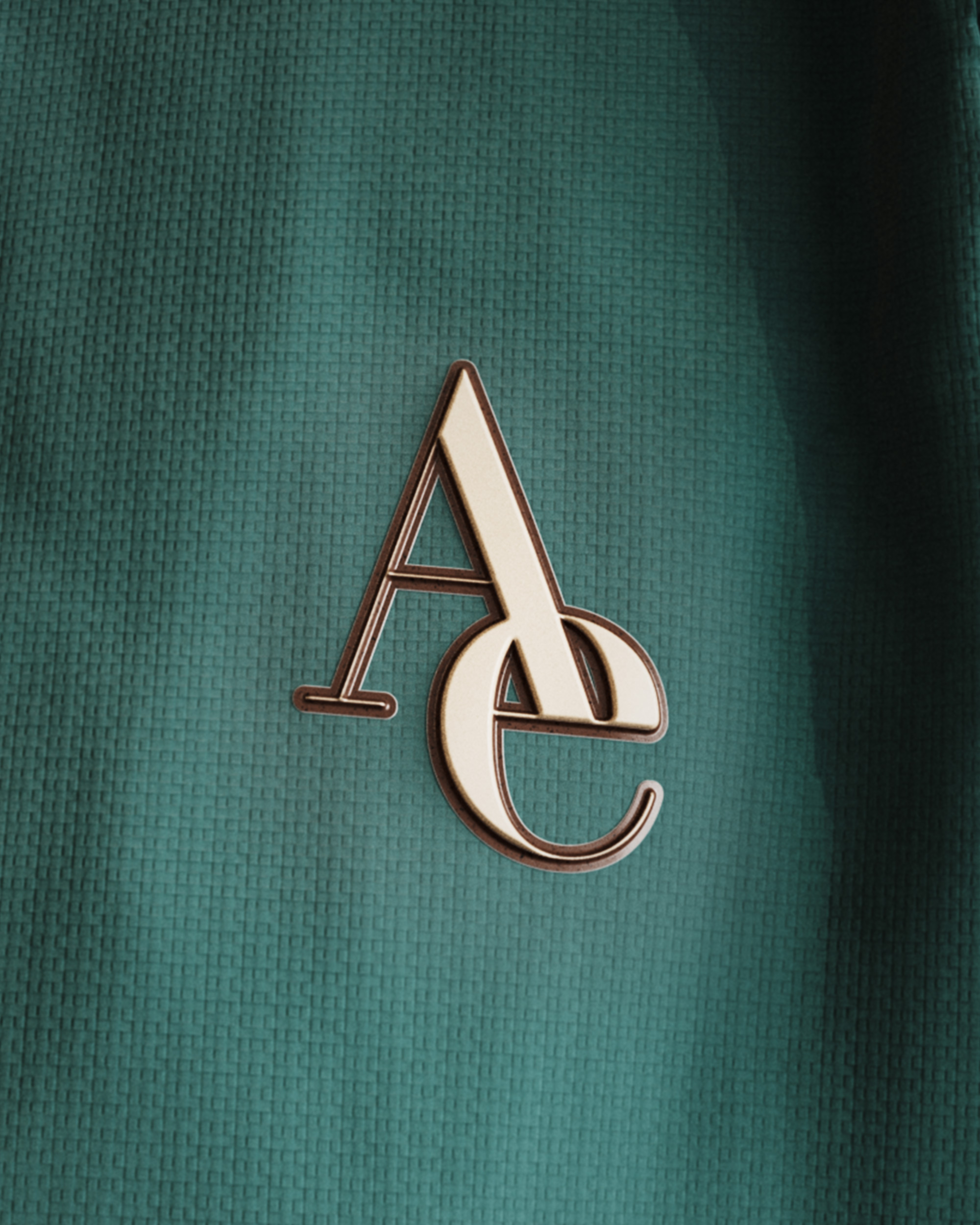 Macro of the Aetheris monogram badge, a cream and brown enamelled A-E ligature raised off green textured piqué, 3D render